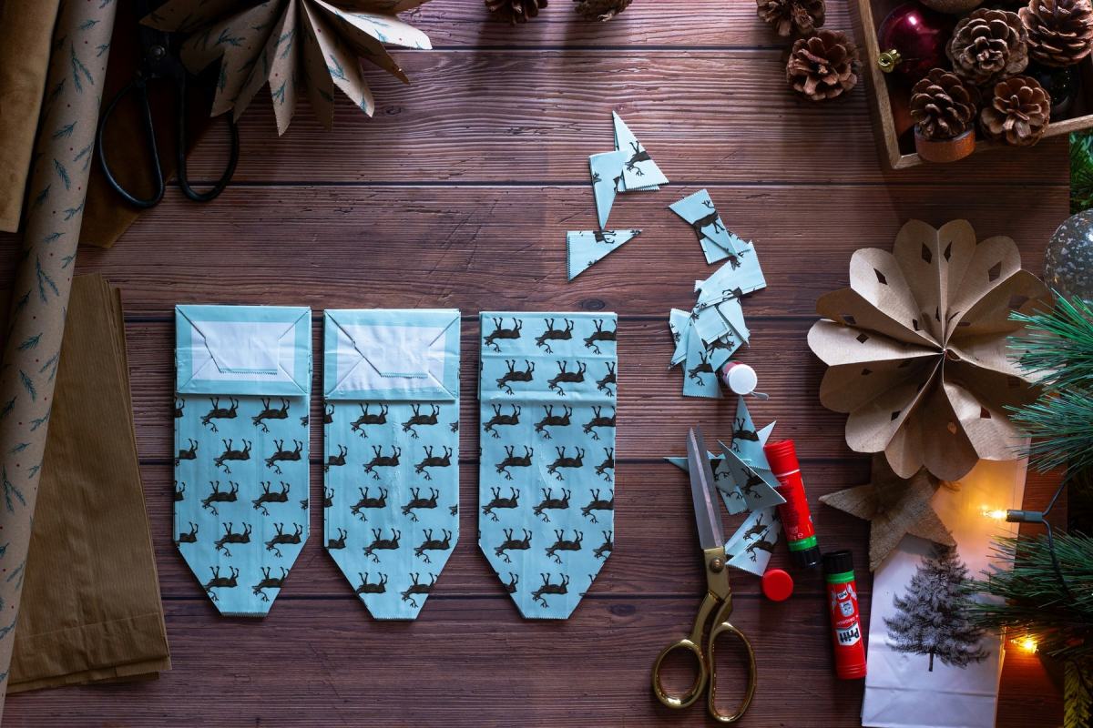 Bastelmaterial für einen Adventskalender: Papiertaschen mit Rentiermuster, Schere und Kleber auf Holz