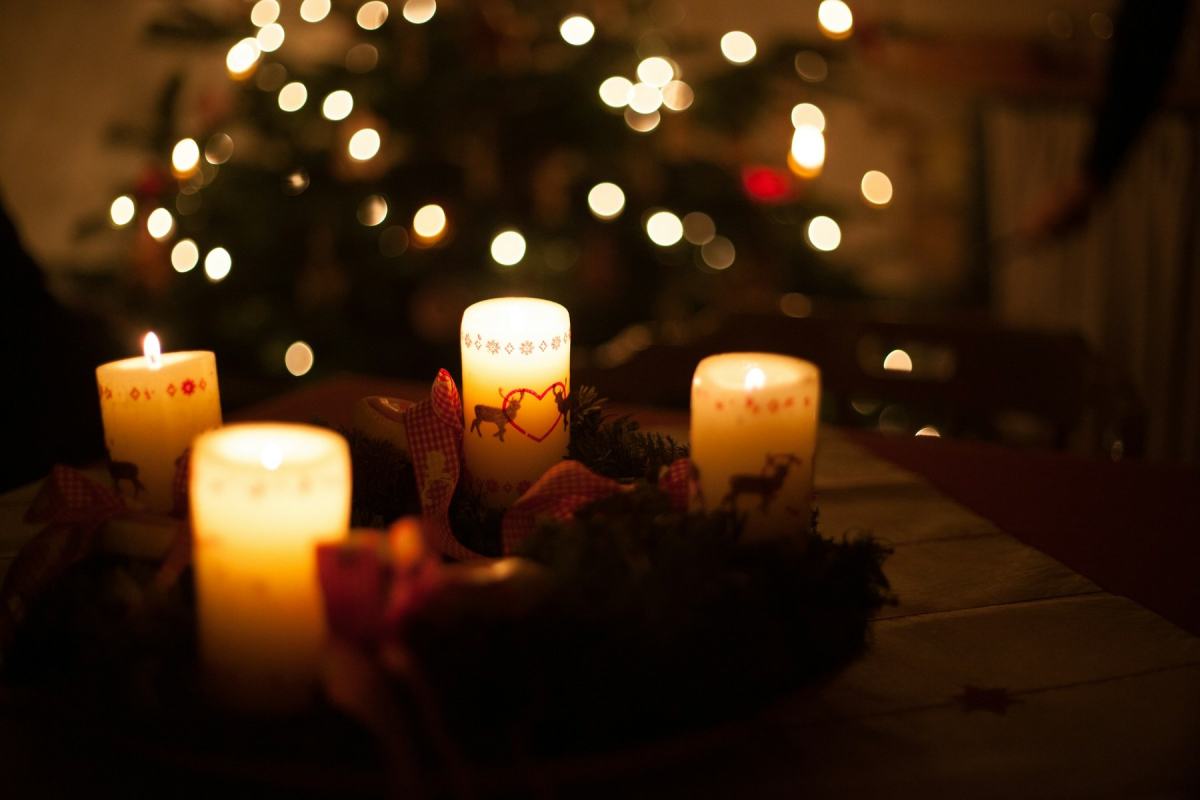 Brennende Kerzen auf einem Adventskranz, dahinter ein Weihnachtsbaum mit Lichterkette