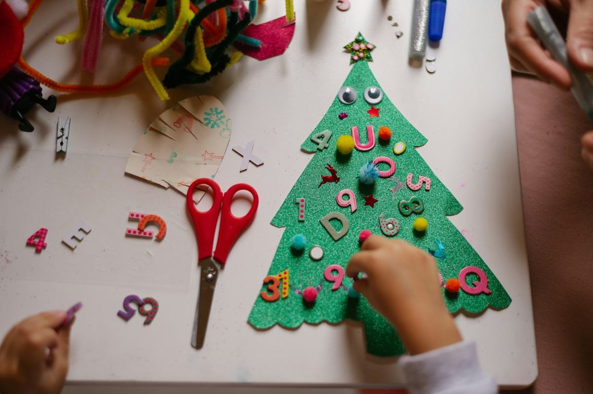 Kinderhände dekorieren einen Filz-Tannenbaum mit bunten Zahlen und Pompons, daneben eine Bastelschere