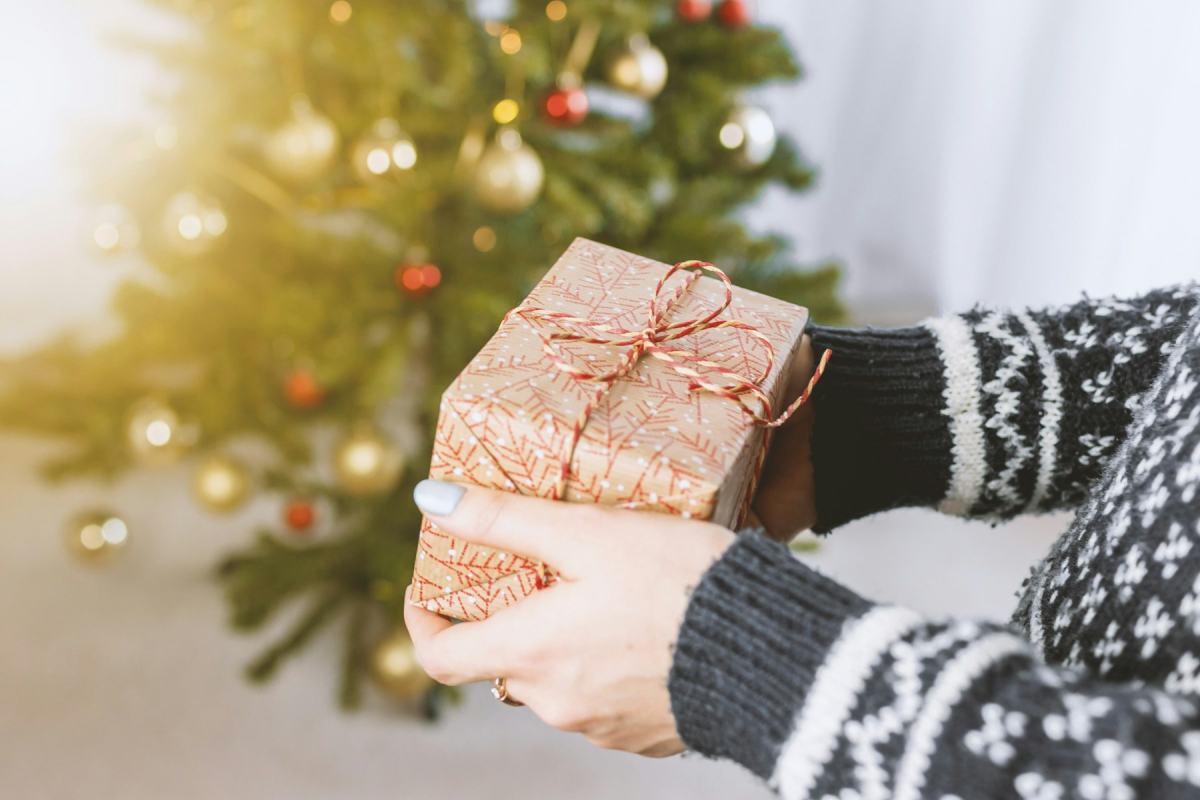 Hände reichen ein verpacktes Geschenk vor einem geschmückten Weihnachtsbaum weiter