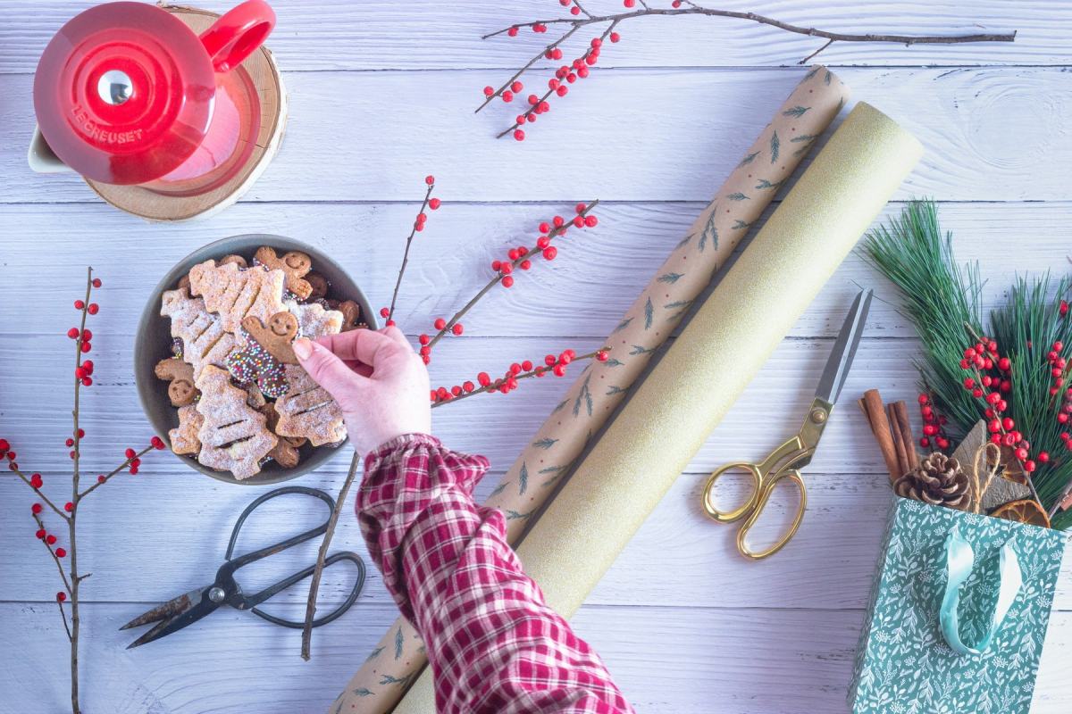 Hände im Karohemd verpacken Geschenke, daneben Plätzchen, Ilexzweige und eine Schere